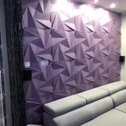 Liliowa ściana z trójwymiarowych paneli geometrycznych, obok szara sofa narożna i okno z zasłonami z delikatnym wzorem.