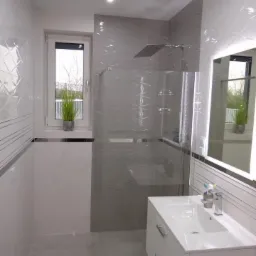 Nowoczesna łazienka z kabiną prysznicową typu walk-in, białą szafką pod umywalką i lustrem z podświetleniem LED. Na ścianach białe i szare płytki z geometrycznym wzorem. W oknie doniczka z zieloną...