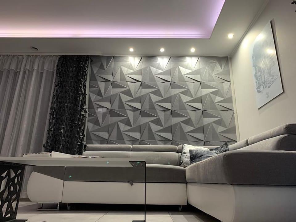 Szara ściana z trójwymiarowych paneli dekoracyjnych w salonie, oświetlenie punktowe sufitowe, narożna sofa z szarymi poduszkami, abstrakcyjny obraz na ścianie, zasłony z dekoracyjną fakturą.