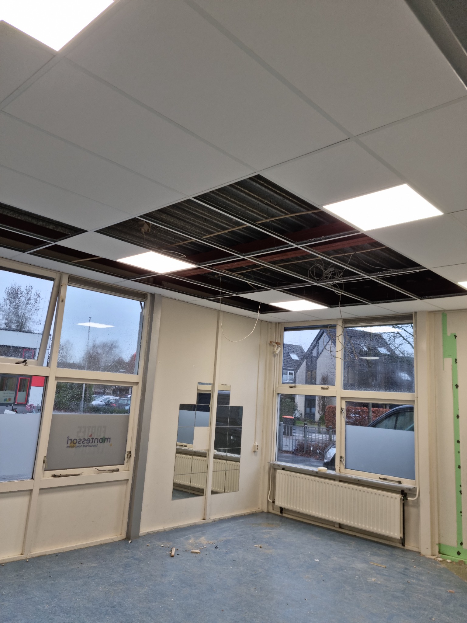 Montaż sufitu podwieszanego kasetonowego w pomieszczeniu biurowym, widoczne częściowo ułożone kasetony, odsłonięta konstrukcja nośna i okablowanie, okna z widokiem na zewnątrz.