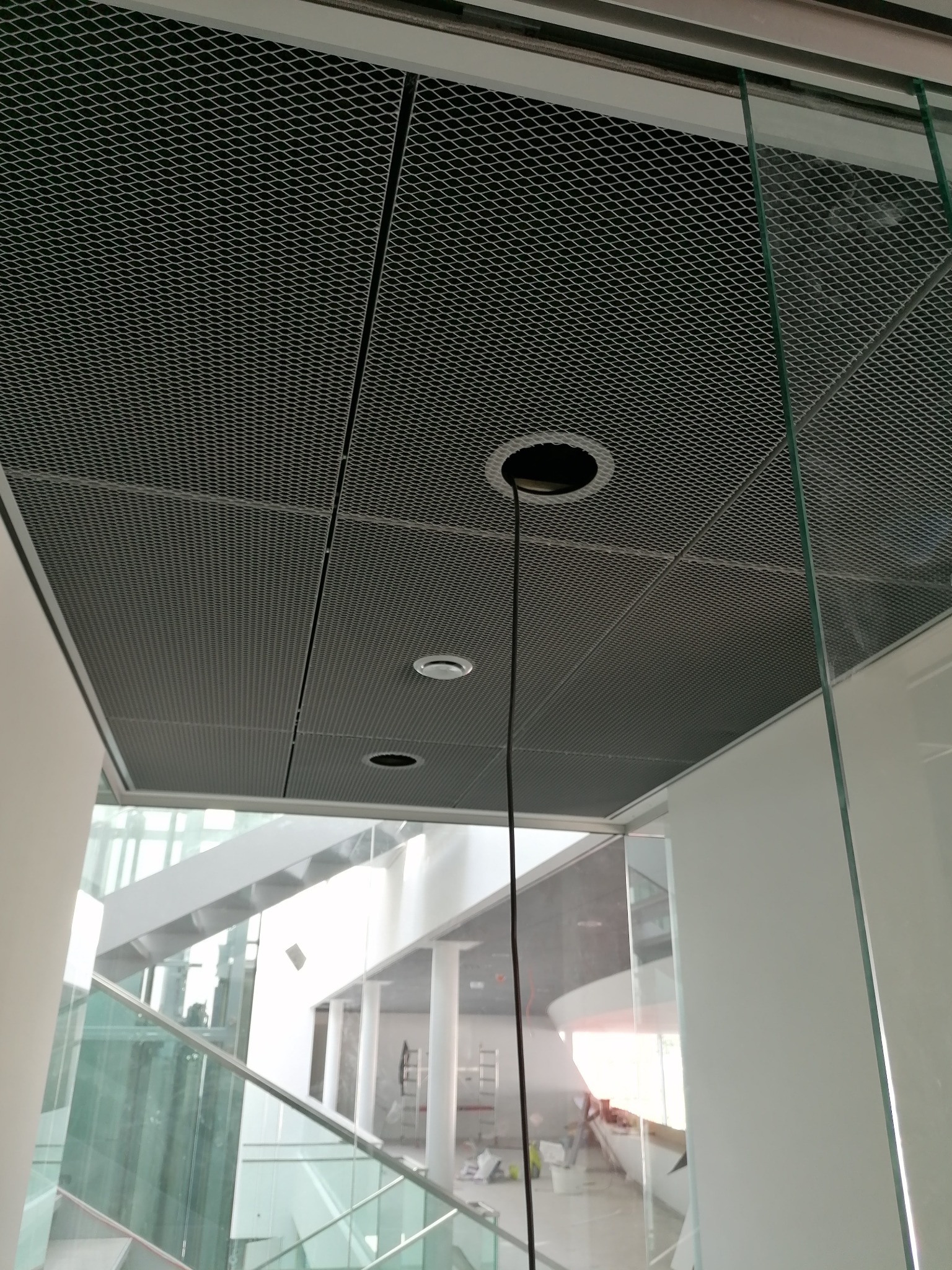 Metalowa, kasetonowa konstrukcja sufitu podwieszanego z widocznymi otworami na oświetlenie i wystającym przewodem elektrycznym, w tle przeszklona klatka schodowa.