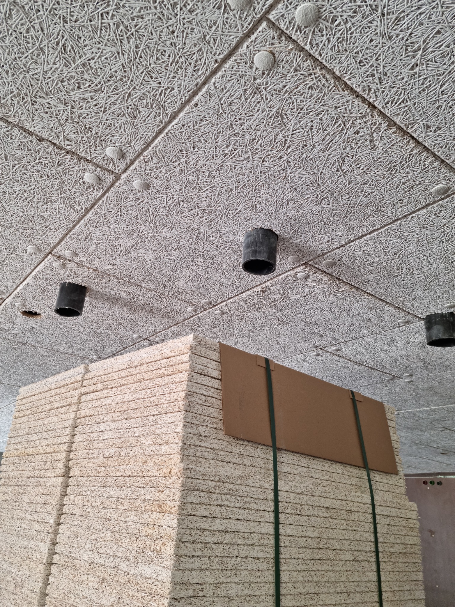 Wykończony sufit z paneli akustycznych z wełny drzewnej, widoczne czarne otwory na oświetlenie i stos paneli gotowych do montażu, zabezpieczony taśmami.