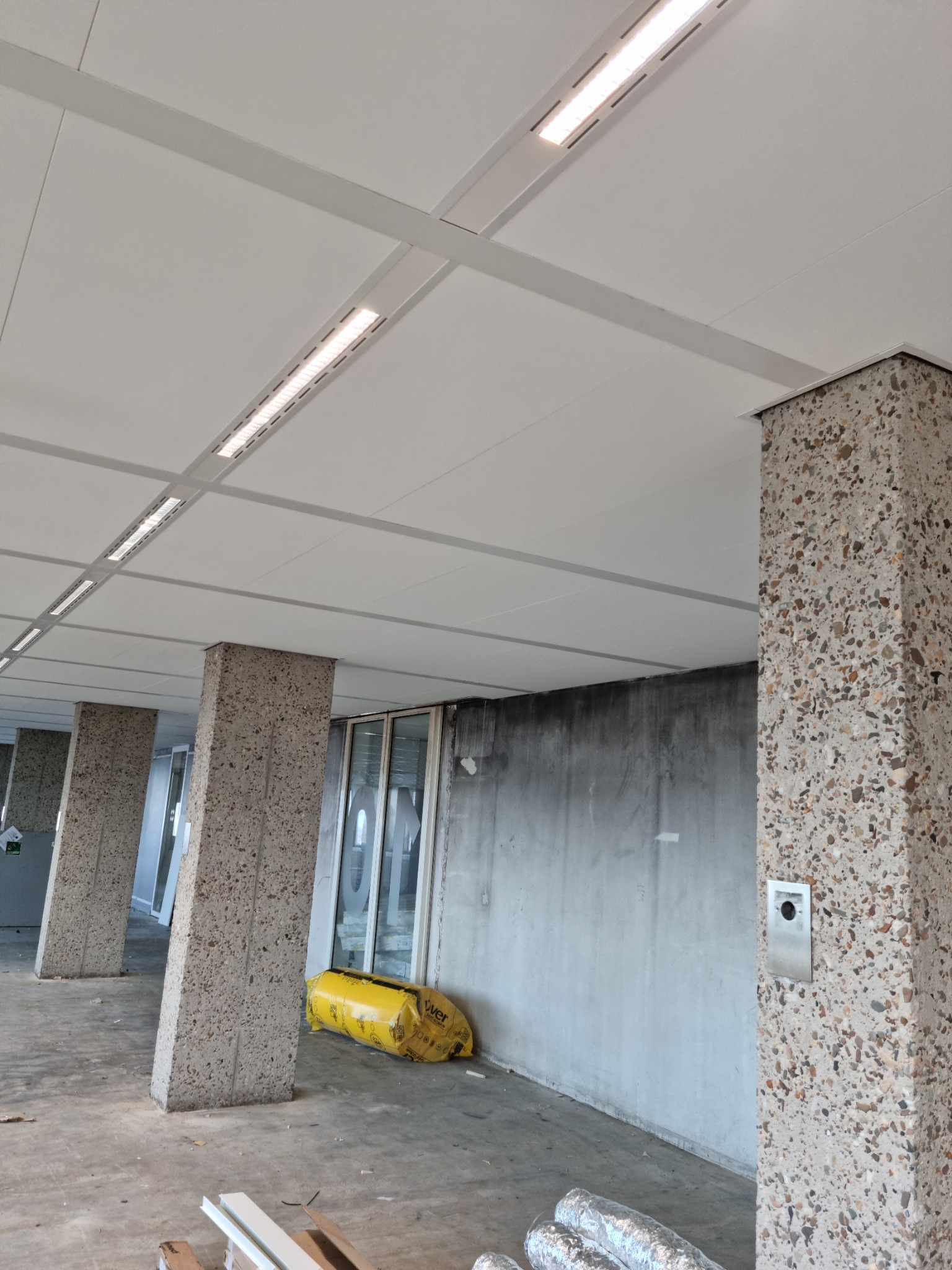 Montaż sufitu kasetonowego z oświetleniem liniowym w przestrzeni komercyjnej, widoczne surowe ściany, kolumny z lastryko i materiały budowlane na podłodze.