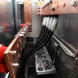 Miedziane szyny zbiorcze w rozdzielnicy elektrycznej, połączone z przewodami oznaczonymi etykietami, widoczne elementy montażowe i podzespoły elektryczne wewnątrz szafy sterowniczej.