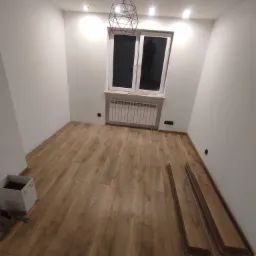 Pokój w trakcie remontu: częściowo ułożona podłoga z paneli, widoczne panele do ułożenia, nowoczesna lampa wisząca i grzejnik pod oknem.