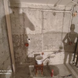 Usługi Elektryczne Radosław Dernałowicz - Surowy stan wnętrza w trakcie prac elektrycznych: widoczne przewody prowadzone po ścianach, puszki instalacyjne, cień osoby pracującej, wiadro na taborecie i butelka płynu na podłodze.