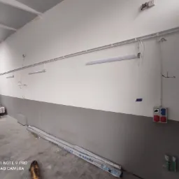 Wnętrze hali w trakcie instalacji oświetlenia: widoczne rury instalacyjne, lampy fluorescencyjne, puszka z gniazdami siłowymi i materiały budowlane na podłodze.