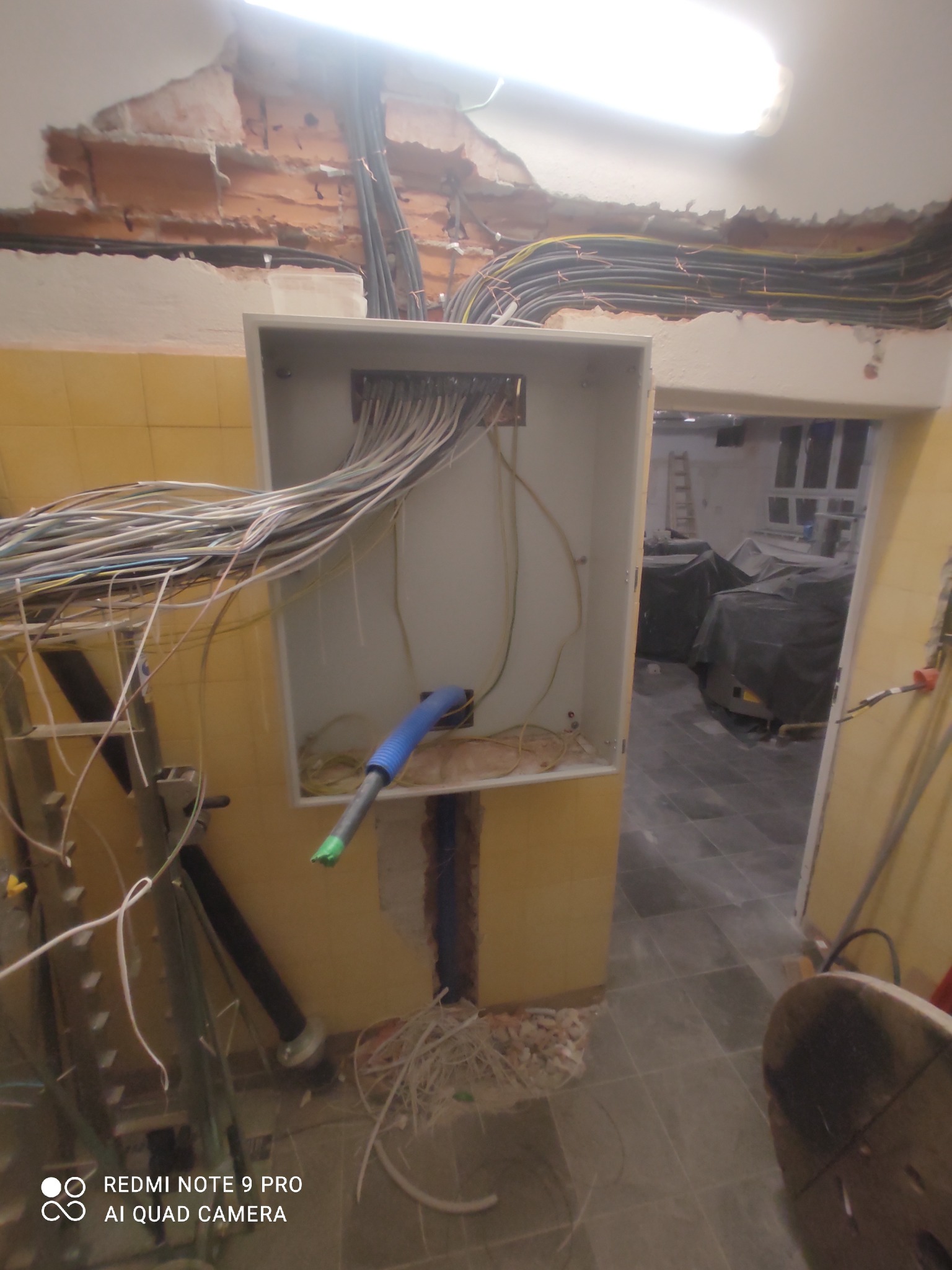 Rozdzielnica elektryczna z licznymi przewodami w trakcie montażu, widoczne kable w ścianie i pod sufitem, fragment ściany z uszkodzonym tynkiem, niebieska rurka karbowana wewnątrz skrzynki.