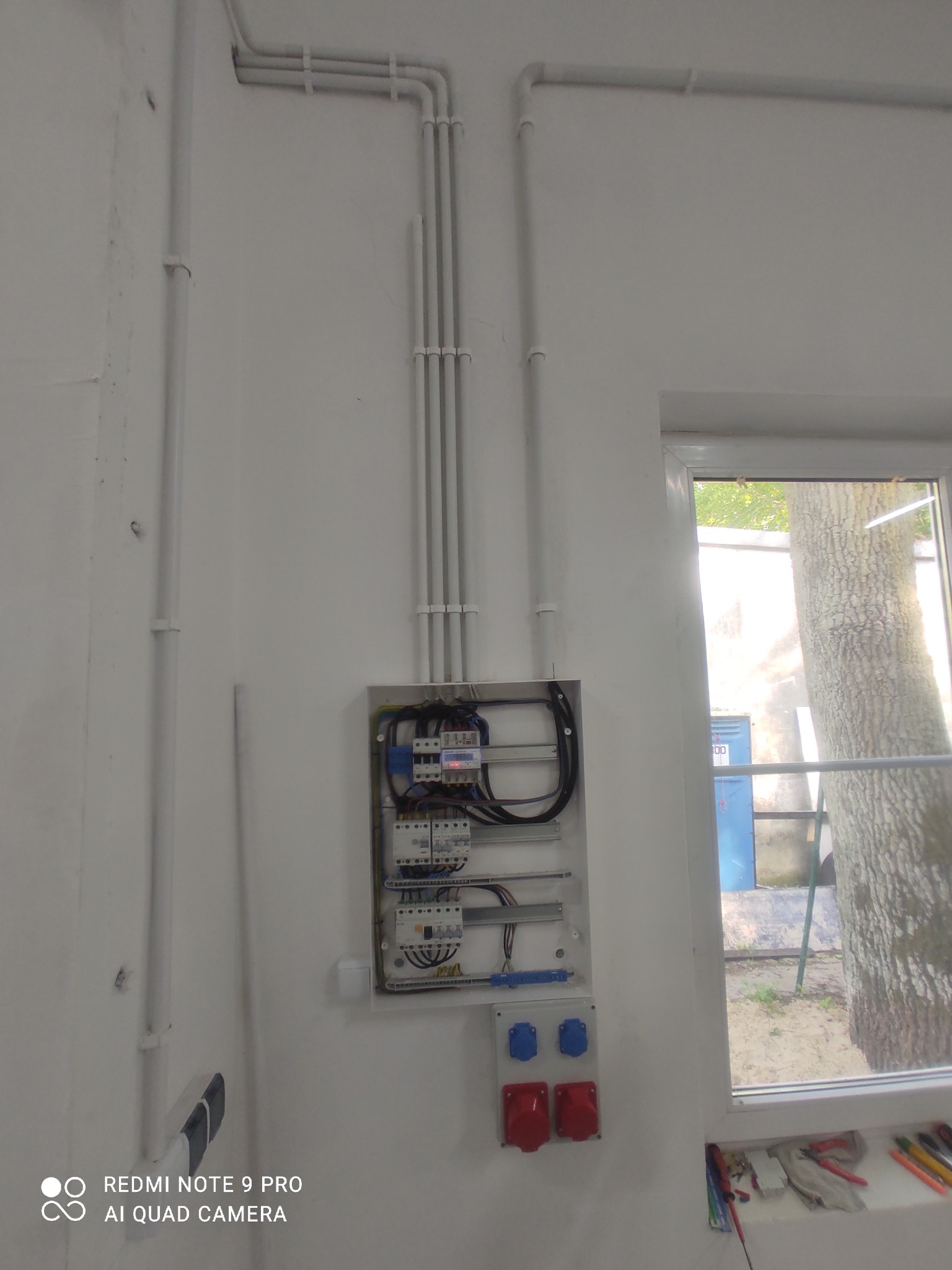 Rozdzielnica elektryczna z zabezpieczeniami, zamontowana na białej ścianie, z widocznymi przewodami w białych rurkach PCV oraz gniazdami siłowymi w kolorze czerwonym i niebieskim. W tle widoczne...