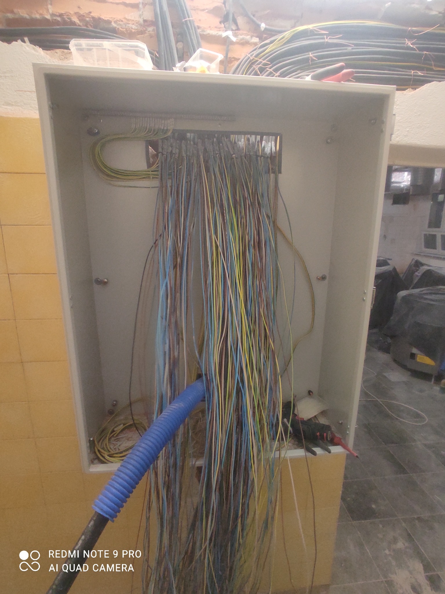 Otwarta skrzynka elektryczna z licznymi przewodami w kolorach niebieskim, żółtym i brązowym, podłączonymi do listwy zaciskowej; widoczny fragment niebieskiego peszla i narzędzia elektryczne.