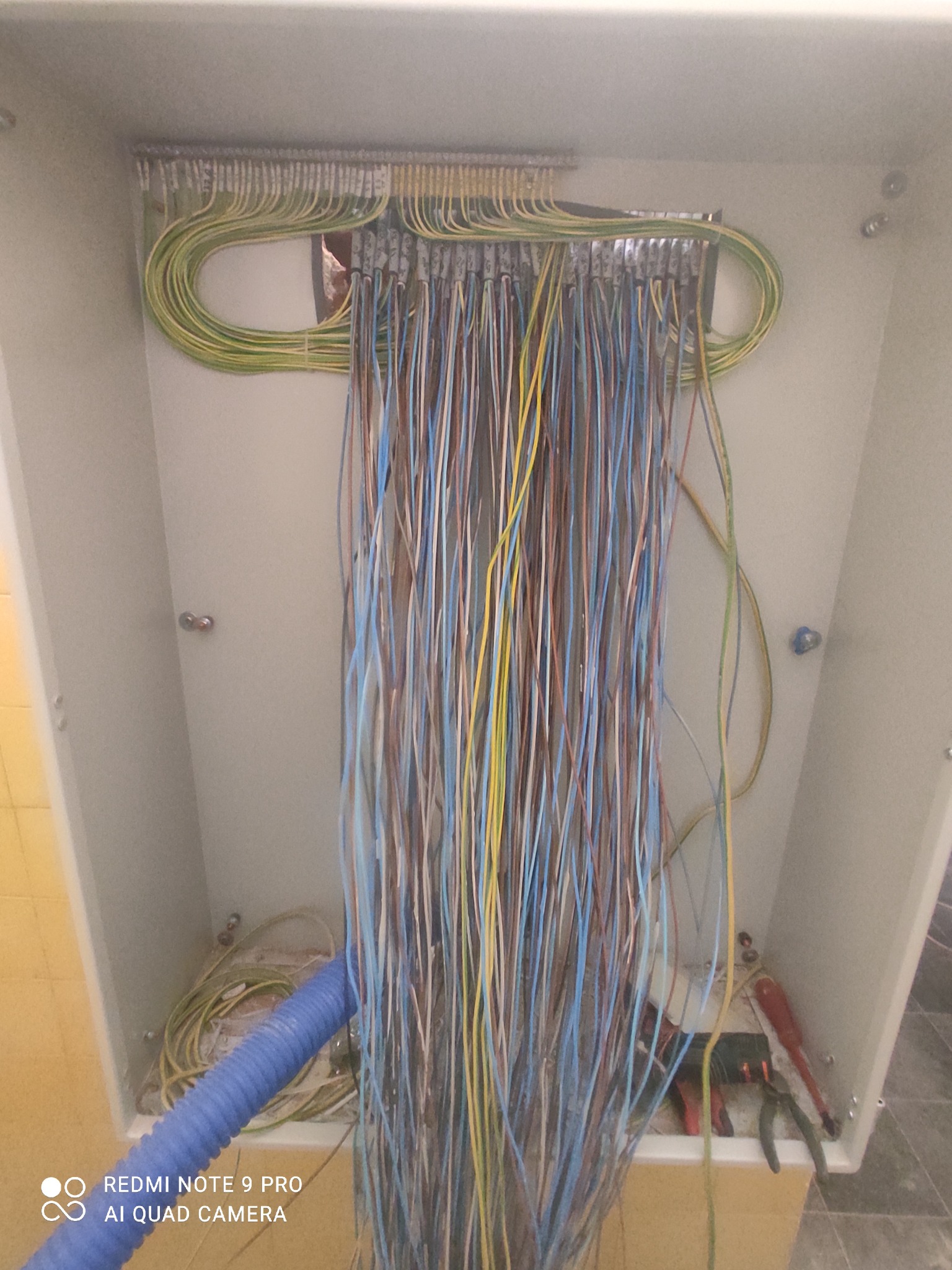Rozdzielnica elektryczna w trakcie montażu: widoczne liczne przewody w różnych kolorach (niebieski, brązowy, żółto-zielony), ułożone w rzędach i podłączone do listew zaciskowych. Na dnie skrzynki...