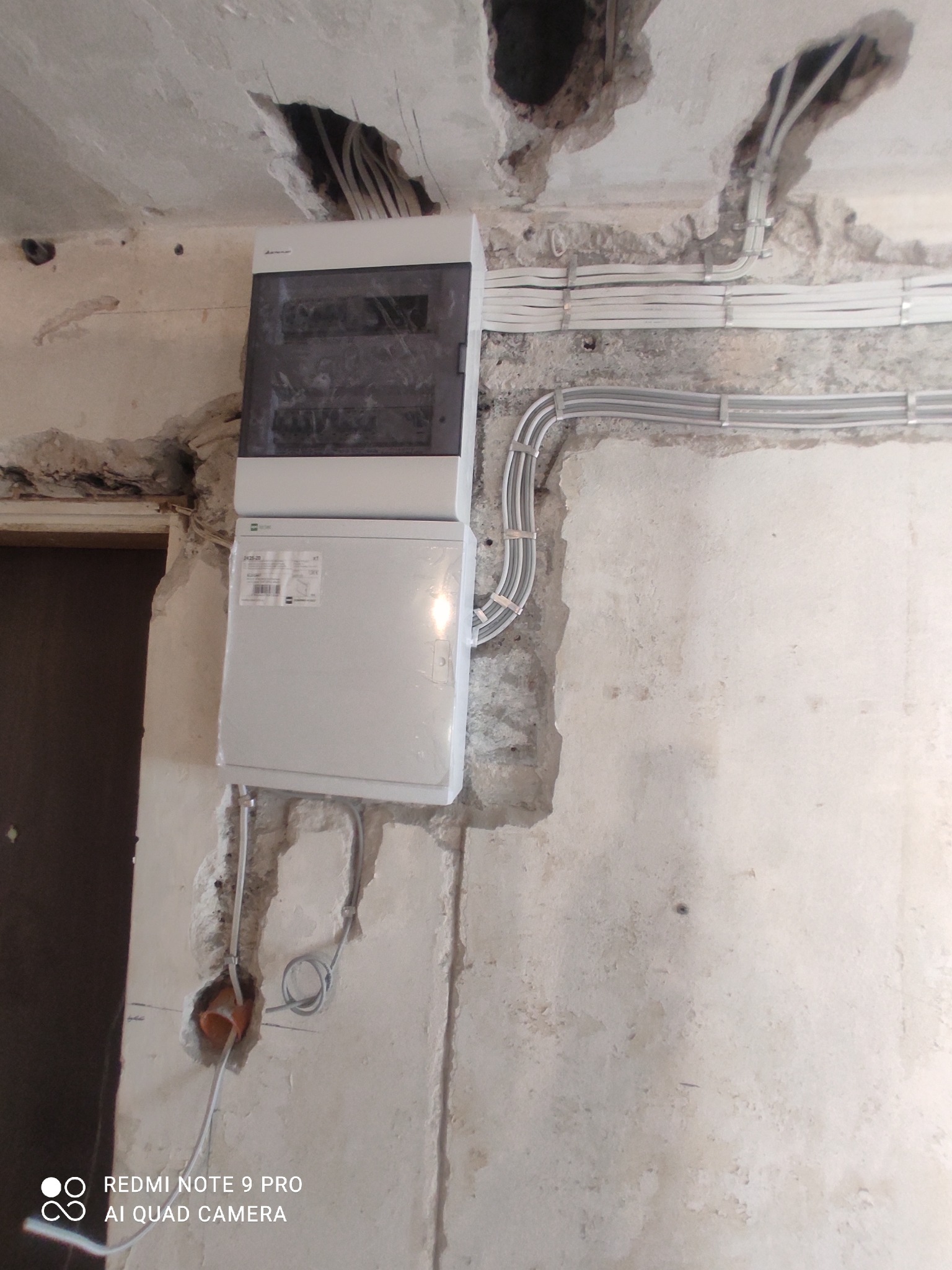 Montaż skrzynki elektrycznej na surowej ścianie, widoczne kable prowadzone wzdłuż ściany i sufitu, ślady po kuciu w tynku.