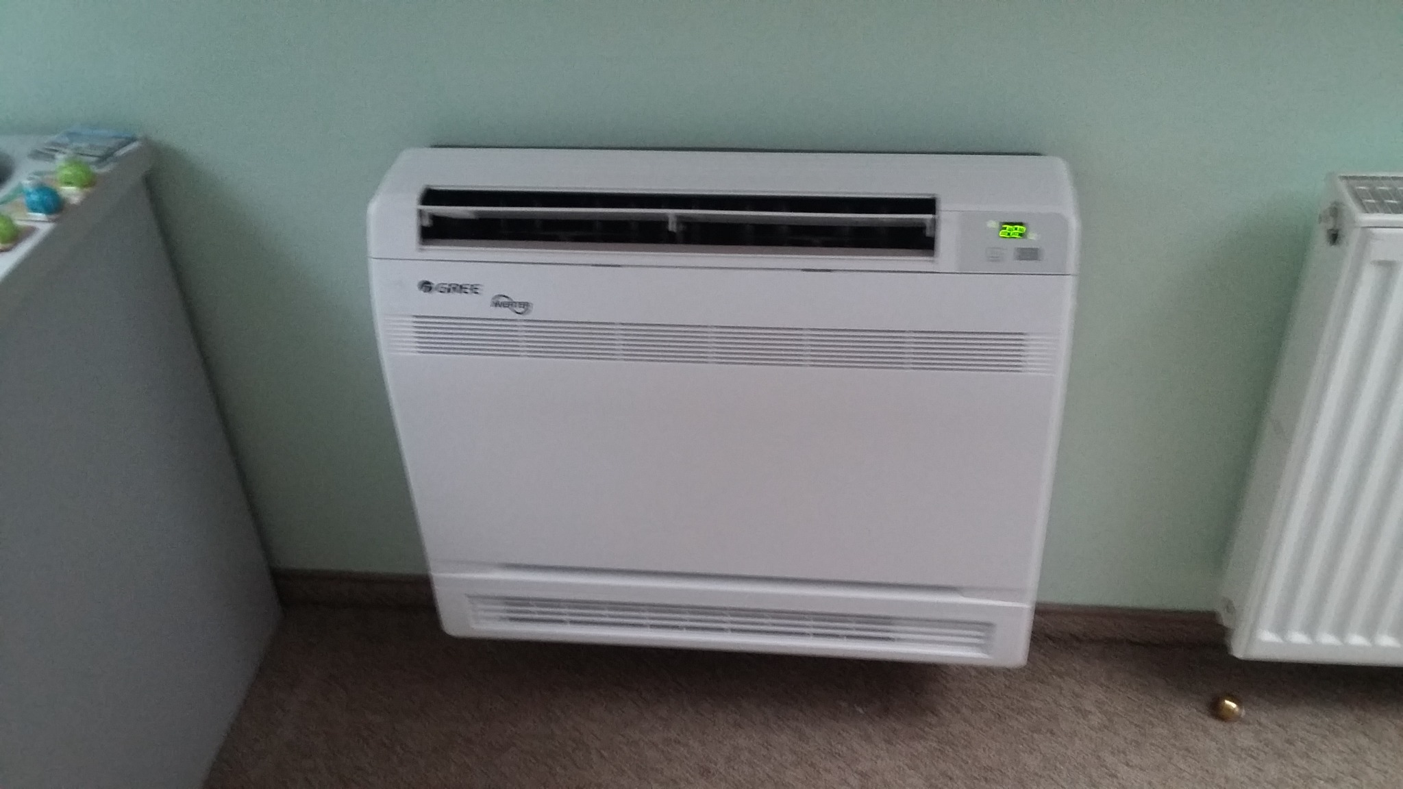 Biały, przypodłogowy klimatyzator marki Gree z wyświetlaczem temperatury, zainstalowany w pomieszczeniu z grzejnikiem.