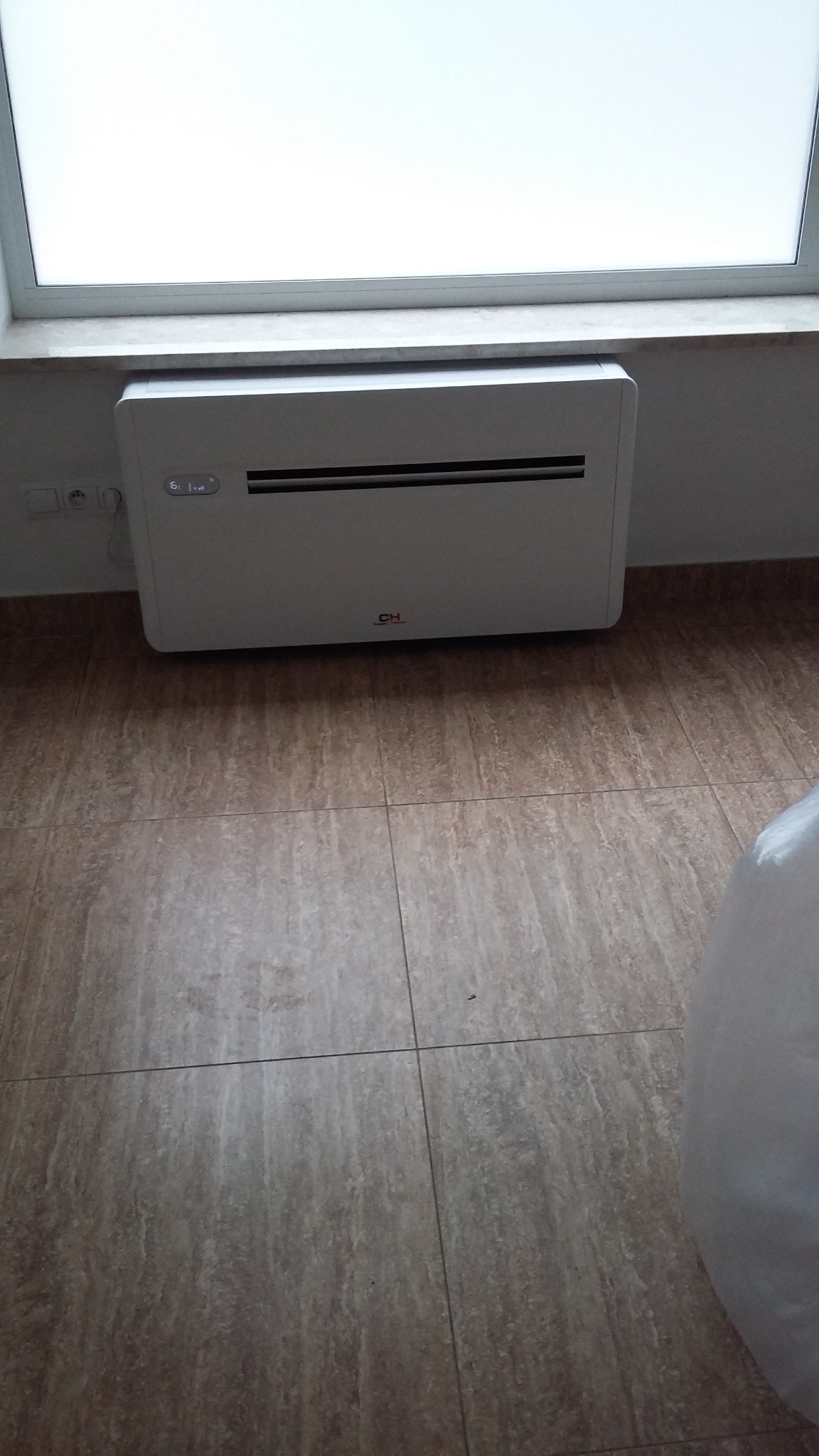 Biały klimatyzator stacjonarny zamontowany pod oknem na tle płytek podłogowych.