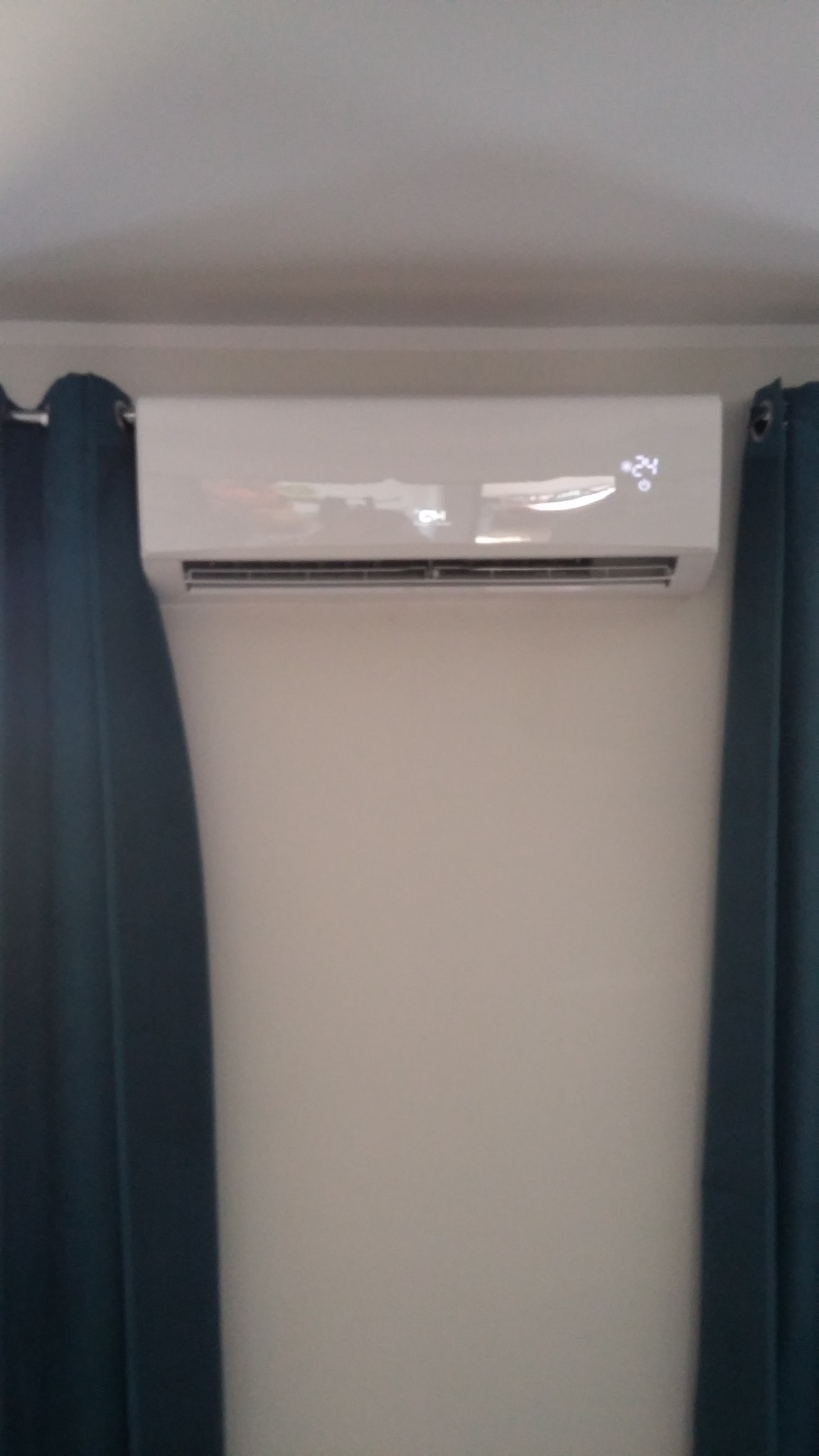 Biały klimatyzator ścienny z wyświetlaczem temperatury 24 stopnie Celsjusza, zawieszony na ścianie między dwoma ciemnozielonymi zasłonami.