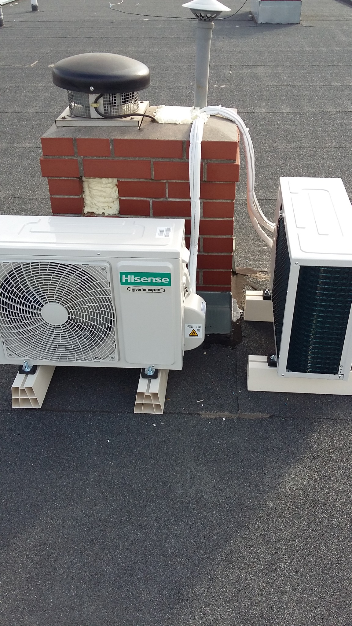 Dwa klimatyzatory Hisense zamontowane na dachu obok ceglanego komina z uszczelnieniami z pianki montażowej.