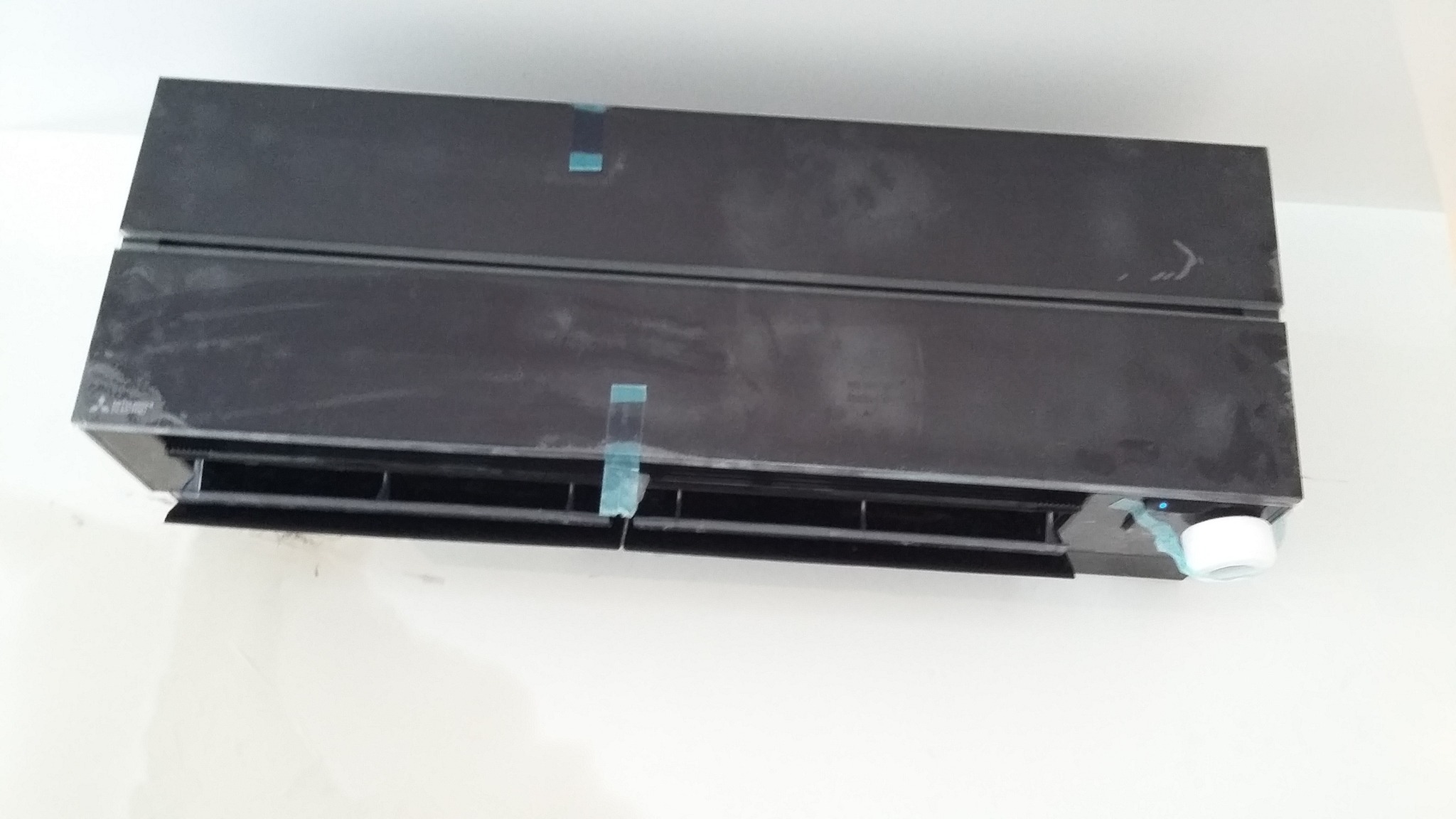 Czarny klimatyzator ścienny, zabezpieczony niebieską taśmą montażową, widoczny na białej ścianie.