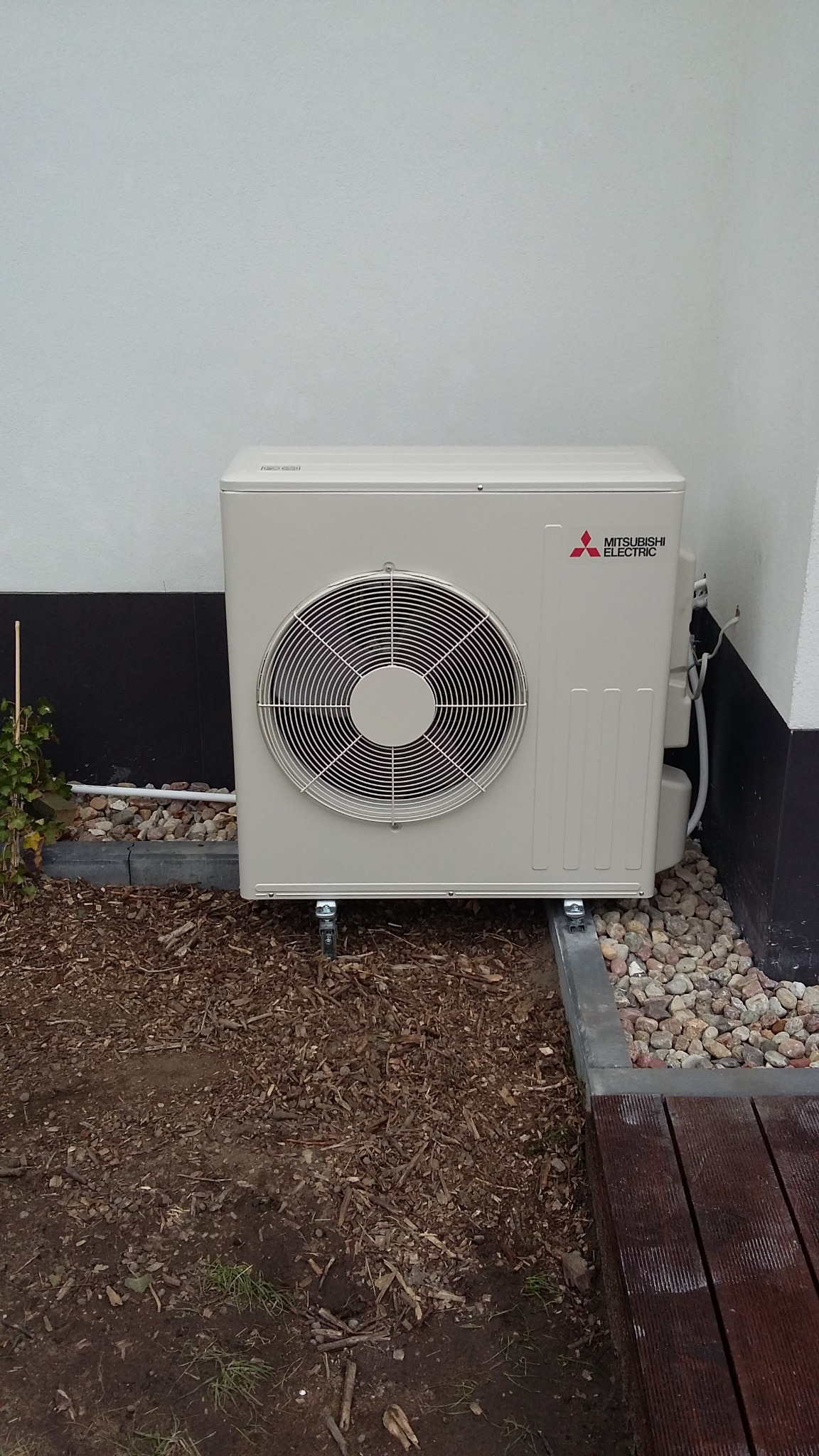 Zewnętrzna jednostka klimatyzacji Mitsubishi Electric na kółkach, umieszczona przy ścianie budynku, otoczona korą i kamieniami ozdobnymi.