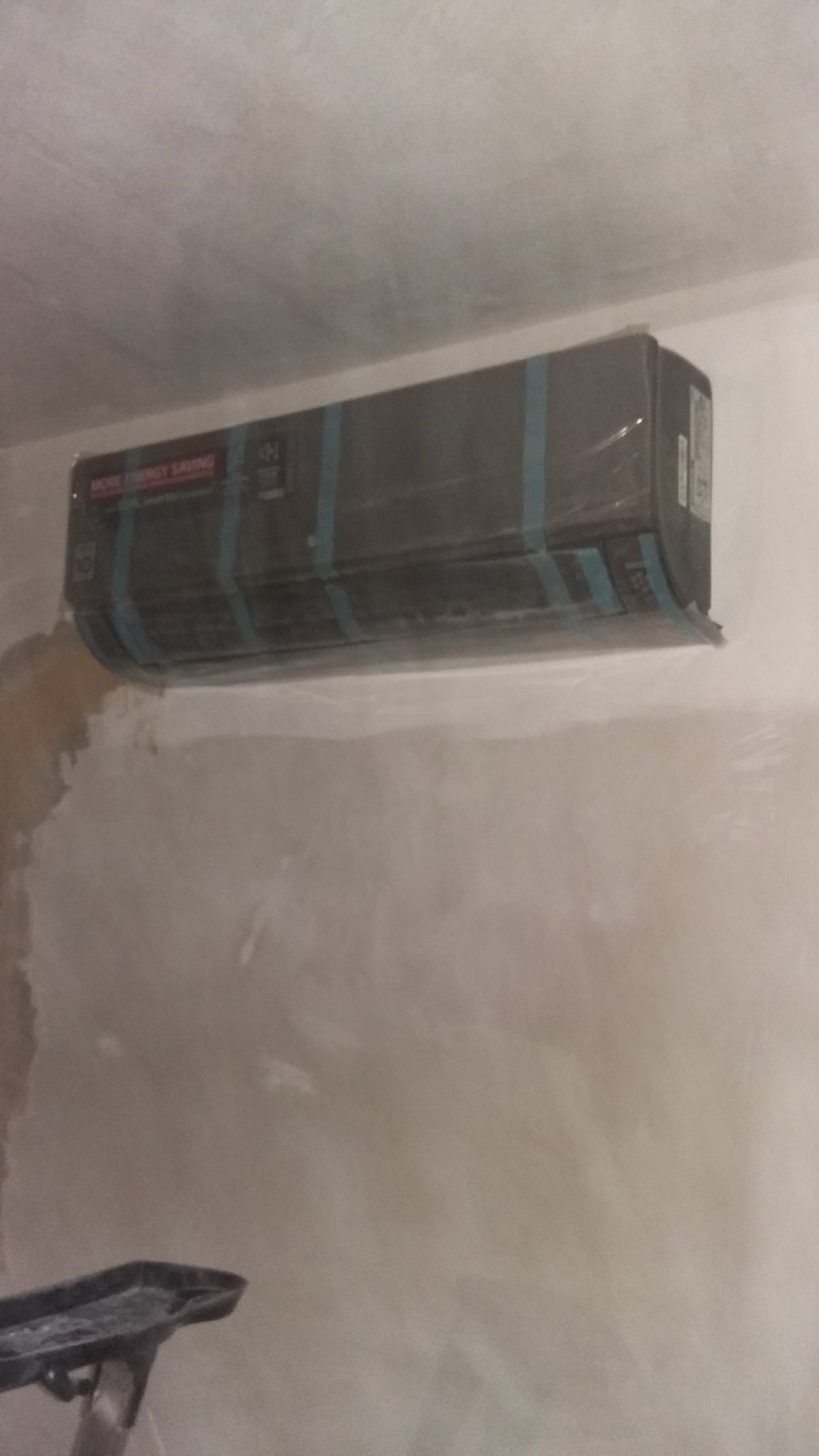 Czarny klimatyzator ścienny, owinięty folią ochronną z niebieską taśmą, zamontowany w surowym wnętrzu podczas remontu.