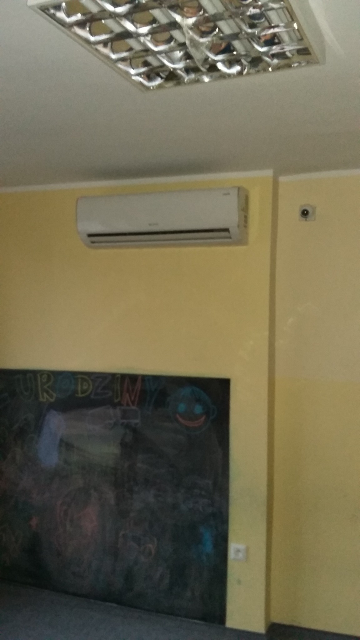 Biały klimatyzator ścienny zamontowany nad tablicą z rysunkami w pomieszczeniu z żółtymi ścianami i kwadratową lampą sufitową.