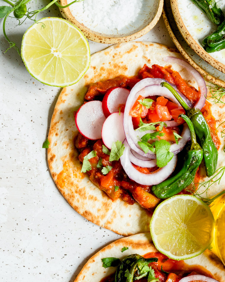 Dwa placki tortilli z farszem, krążkami cebuli, rzodkiewką, papryczkami padrón i ćwiartkami limonki, ujęcie z góry na jasnym tle.