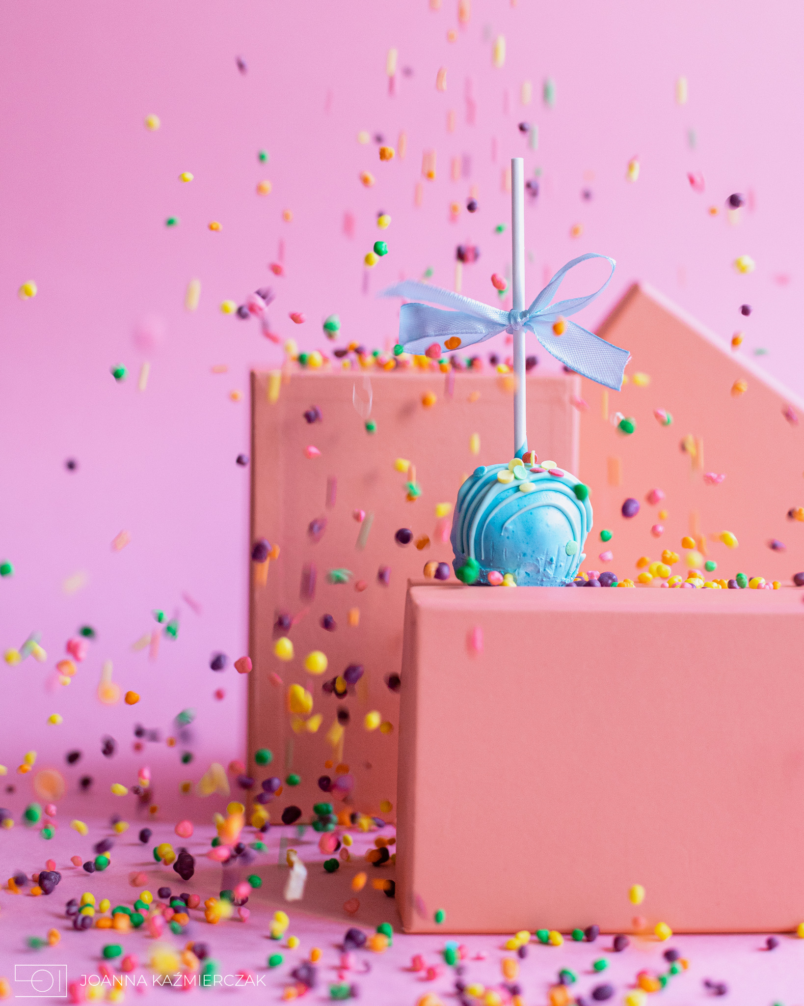 Niebieski cake pop z wstążką i kolorowymi posypkami na różowym tle i różowych blokach.