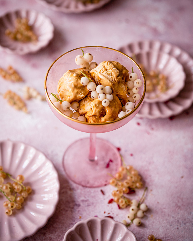 Kremowe lody w różowym kieliszku z obwódką, udekorowane białymi porzeczkami na tle różowych talerzyków i blatu.