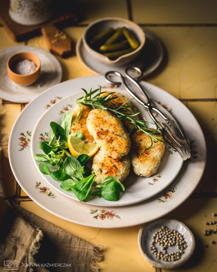 Kulinarna fotografia: Kotlety rybne na talerzu z motywem kwiatowym, udekorowane roszponką, cytryną i rozmarynem, obok szczypce, marynowane ogórki, sól i ziarna pieprzu na żółtej, kafelkowej...
