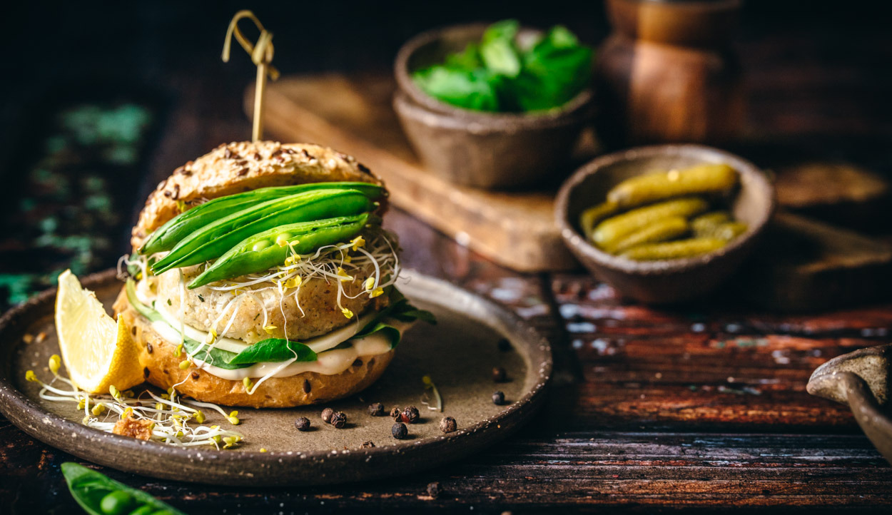 Stylizowany wegański burger z groszkiem cukrowym, kiełkami i cytryną na rustykalnym talerzu i drewnianym tle.