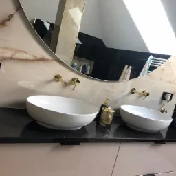 Łazienka z dwoma białymi umywalkami nablatowymi na czarnym, błyszczącym blacie z drobinkami, złota armatura, duże okrągłe lustro z oświetleniem odbijającym się w tafli, beżowe marmurowe ściany.