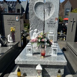 Granis Kamieniarstwo - Granitowy pomnik z wyciętym sercem i rzeźbą anioła, ozdobiony kwiatami i lampionami, na tle zabudowań mieszkalnych z panelami słonecznymi.