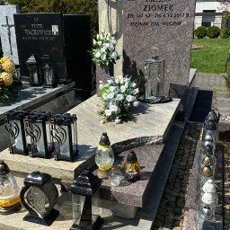 Granis Kamieniarstwo - Granitowy nagrobek z dwoma tablicami, kwiatami i lampionami na cmentarzu, widoczne inne groby w tle.