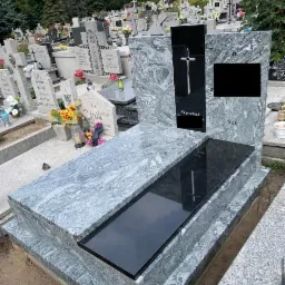 Nowoczesny nagrobek z szarego granitu z czarnym elementem centralnym z krzyżem, widok na cmentarz z licznymi nagrobkami w tle.