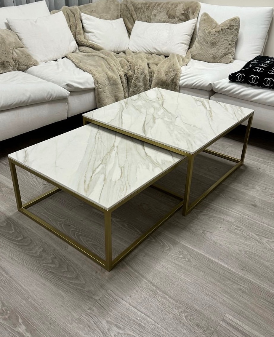 Dwa stoliki kawowe z blatami imitującymi marmur i złotymi ramami, ustawione jeden pod drugim, na tle białej sofy z poduszkami i beżowym pledem.