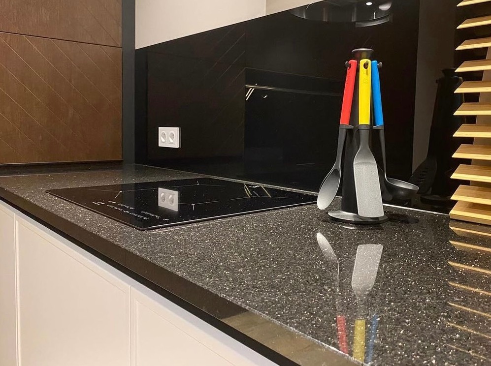 Błyszczący blat kamienny w kuchni z płytą indukcyjną i zestawem kolorowych przyborów kuchennych na tle czarnego panelu ściennego z subtelnym wzorem jodełki.