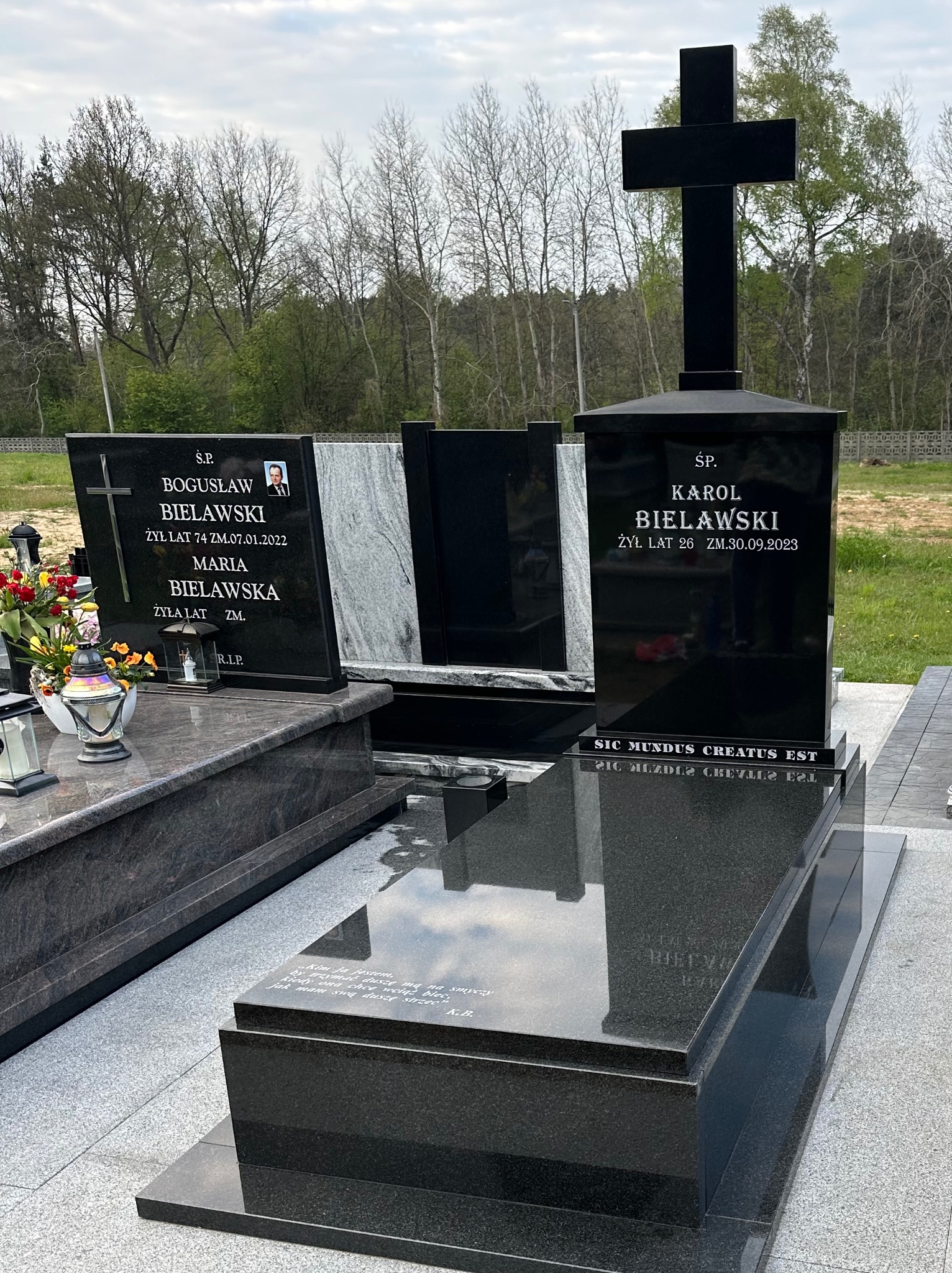 Czarny, podwójny pomnik z krzyżem na cmentarzu, z inskrypcjami i zdjęciami zmarłych, otoczony drzewami i zielenią.