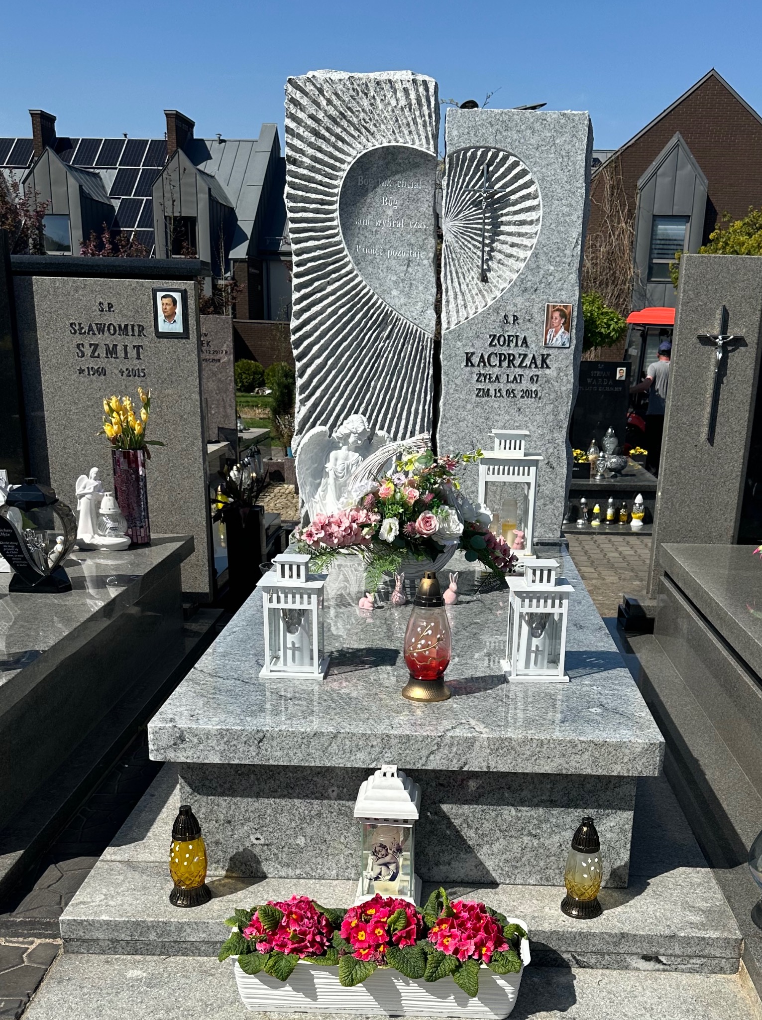 Granitowy pomnik z wyciętym sercem i rzeźbą anioła, ozdobiony kwiatami i lampionami, na tle zabudowań mieszkalnych z panelami słonecznymi.