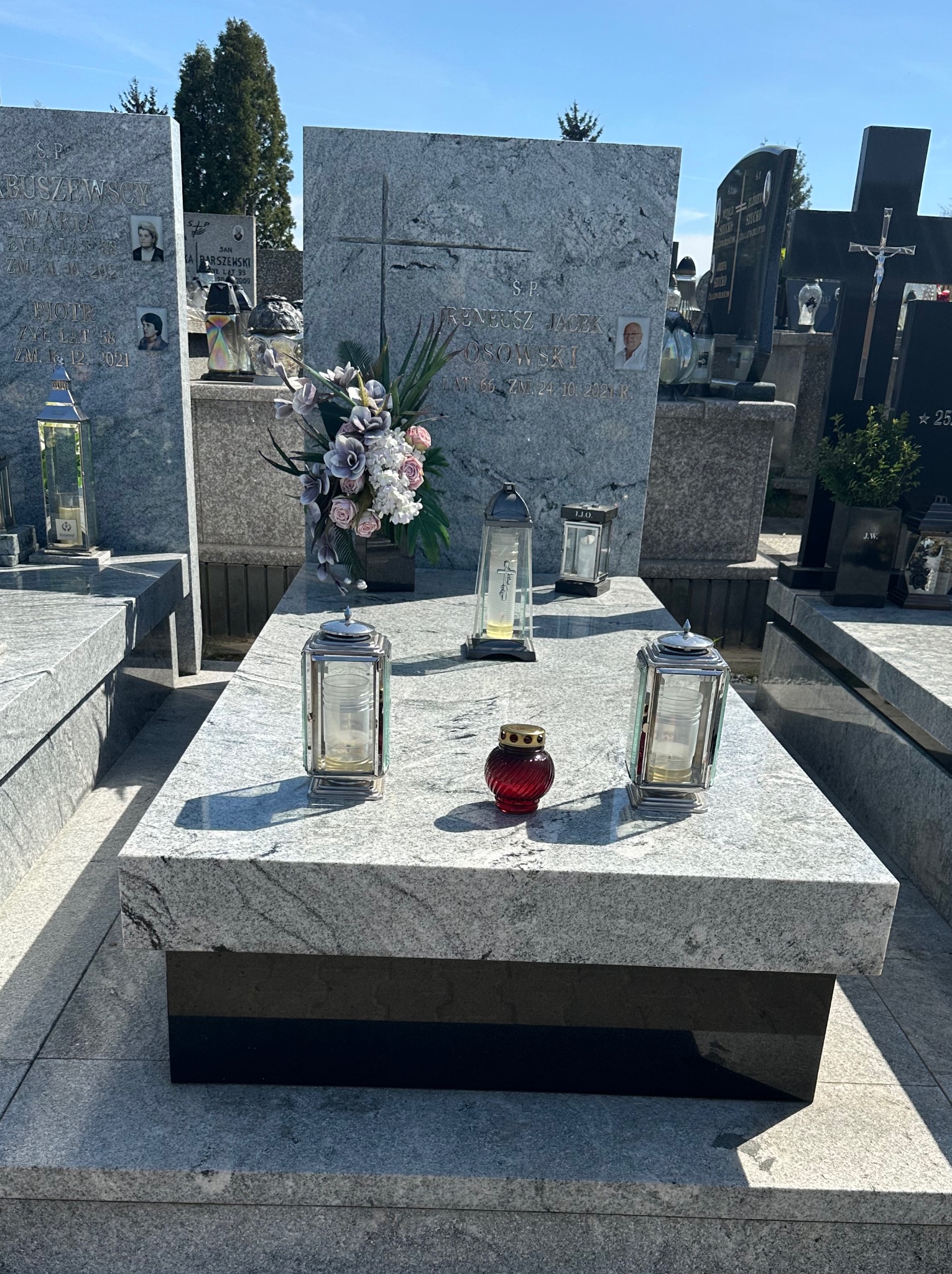 Jasnoszary, granitowy nagrobek z czarnym cokołem, ozdobiony kwiatami w wazonie, dwoma lampionami i czerwoną zniczem, na tle cmentarza w słoneczny dzień.