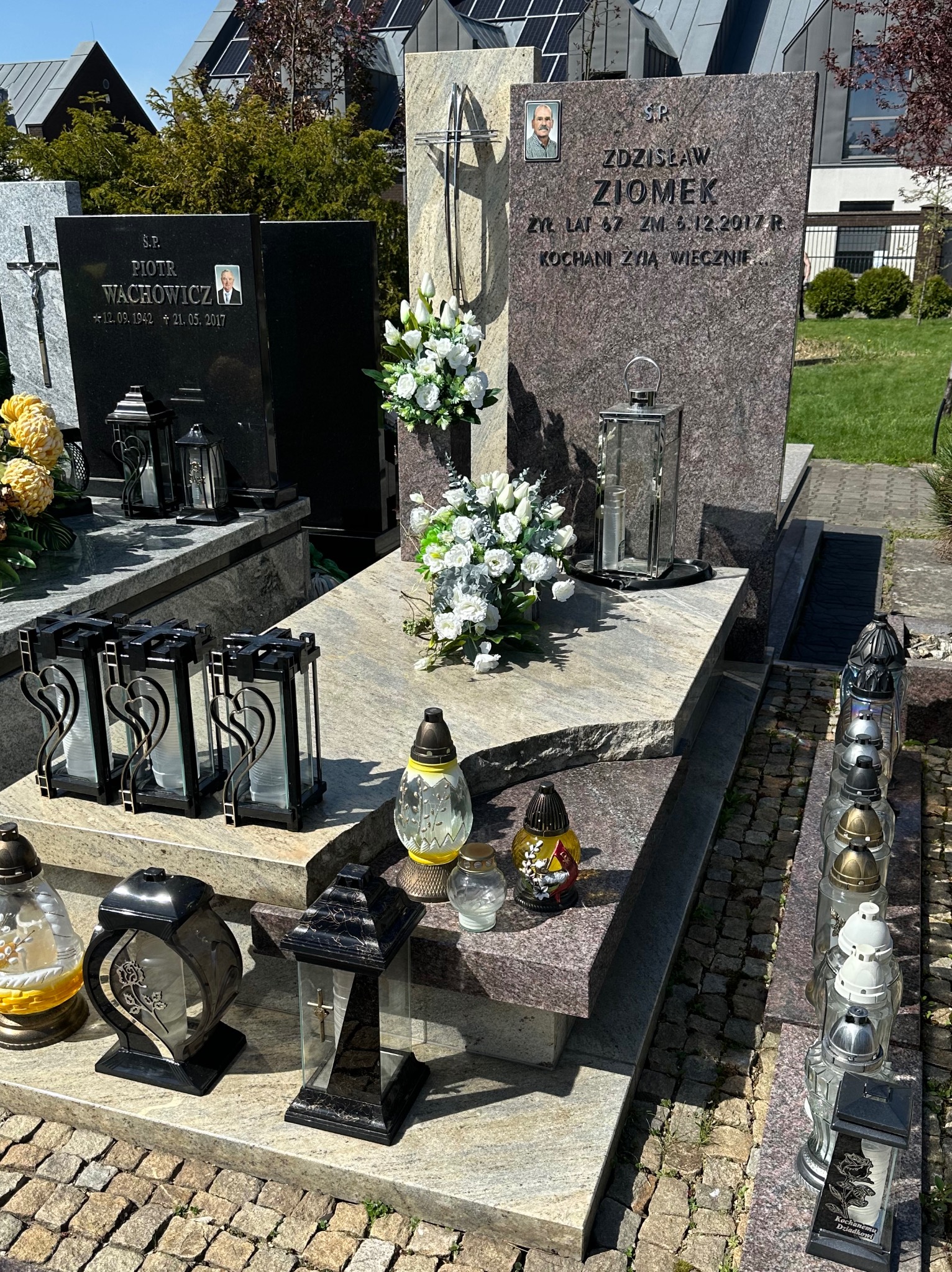 Granitowy nagrobek z dwoma tablicami, kwiatami i lampionami na cmentarzu, widoczne inne groby w tle.