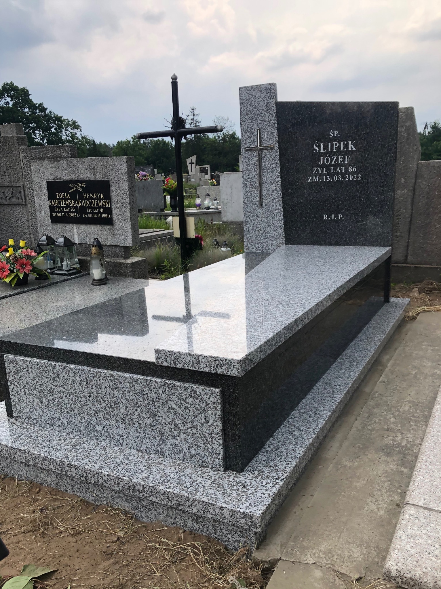 Granitowy nagrobek z krzyżem, polerowany blat, inskrypcje z imieniem i datami, otoczenie cmentarza z innymi grobami i zielenią.