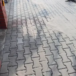 Ułożona szara kostka brukowa typu I z widocznymi śladami piasku na powierzchni, częściowo oczyszczona, z widocznymi elementami ogrodzenia i fragmentem ściany budynku w tle.