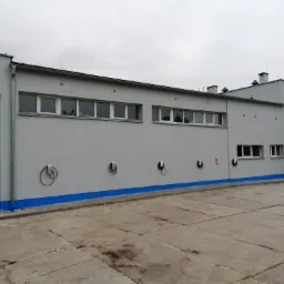 Szary budynek przemysłowy z niebieskimi akcentami, widoczne okna, rury spustowe i punkty ładowania pojazdów elektrycznych na elewacji.