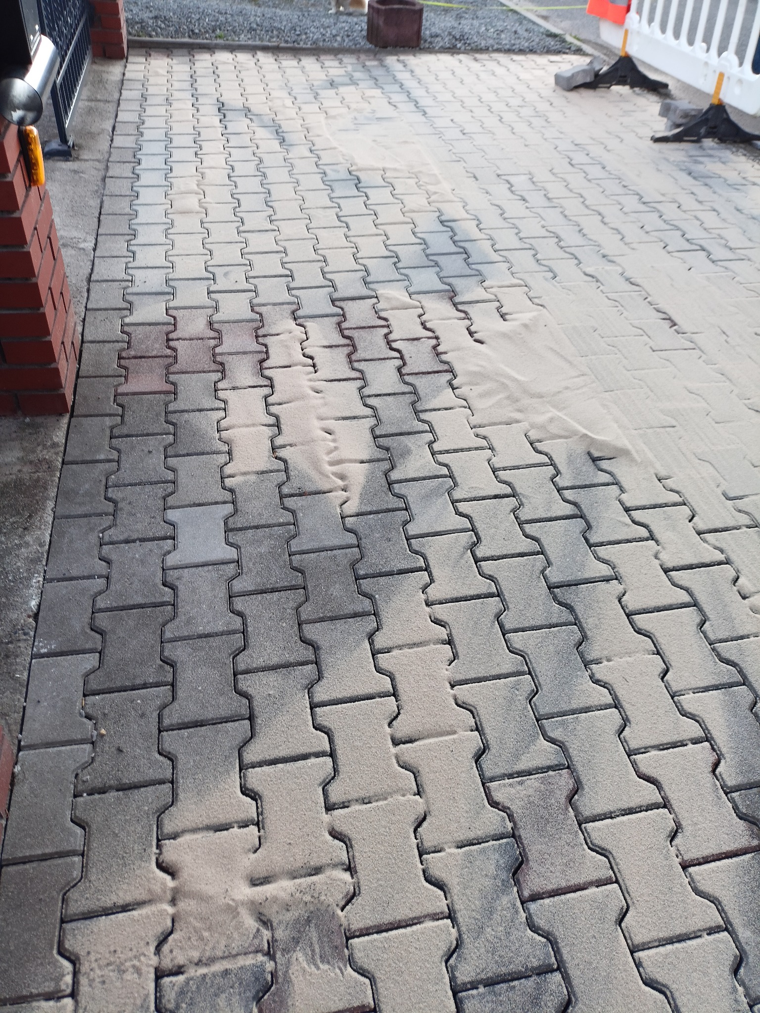 Ułożona szara kostka brukowa typu I z widocznymi śladami piasku na powierzchni, częściowo oczyszczona, z widocznymi elementami ogrodzenia i fragmentem ściany budynku w tle.