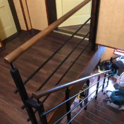 Nowoczesne schody z drewnianą poręczą i metalowymi szczeblami, widok z góry na brązowe stopnie i podłogę imitującą drewno, w tle widoczny wózek dziecięcy.