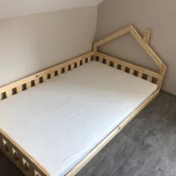 Drewniane łóżko dziecięce w kształcie domku z białym materacem, widok z góry, surowe wykończenie drewna, szare panele podłogowe.