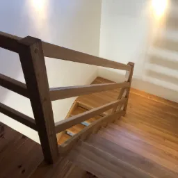 Klatka schodowa z drewnianymi schodami i prostą, drewnianą poręczą z trzema poziomami, widok z góry, ciepłe oświetlenie podkreśla naturalny odcień drewna.