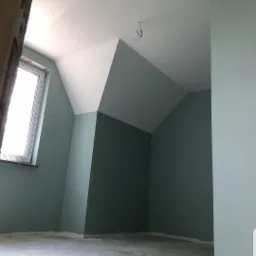 Pokój na poddaszu po malowaniu ścian na odcień zieleni, z widocznym oknem dachowym i kablem zwisającym z sufitu; surowa podłoga przed wykończeniem.