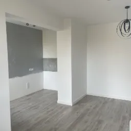 Wnętrze pokoju z szarą ścianą częściowo wykończoną, drewnopodobną podłogą, białymi ścianami i nowoczesnymi lampami wiszącymi.