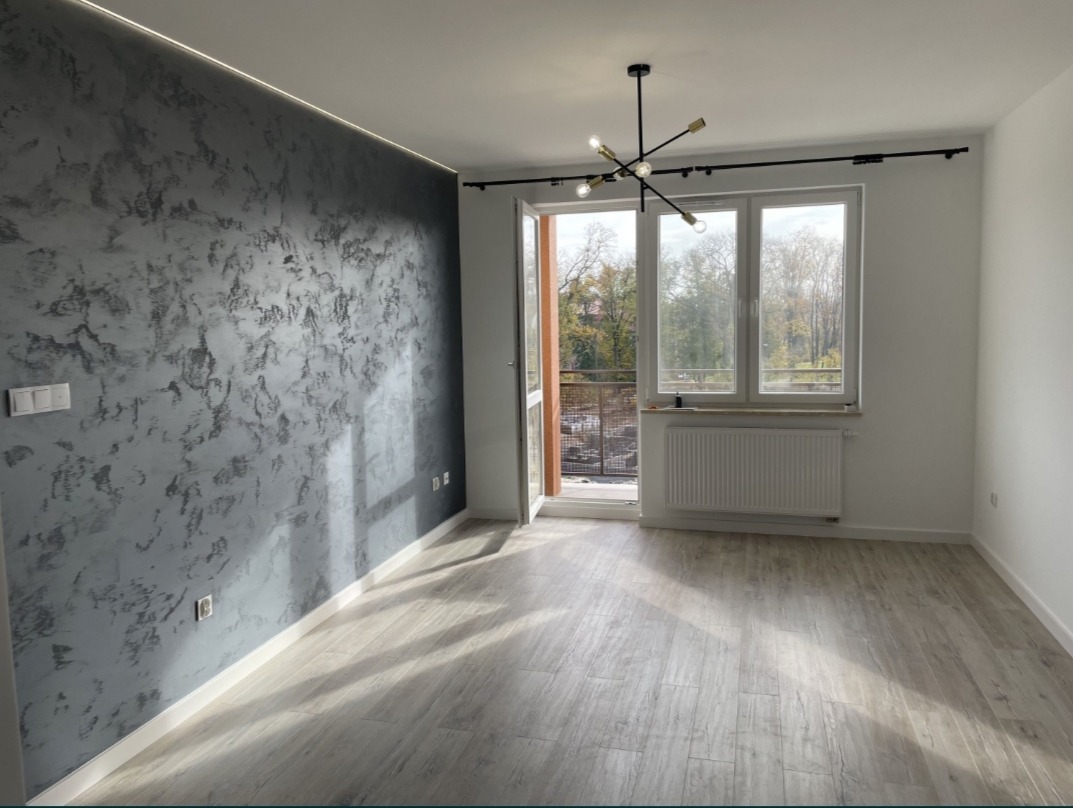 Pusty pokój po remoncie z dekoracyjną, szarą ścianą z efektem betonu, drewnopodobną podłogą, nowoczesną lampą sufitową i oknem z widokiem na drzewa.