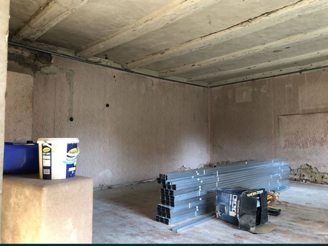 Wnętrze w trakcie prac remontowych: widoczne surowe ściany z resztkami tapety, betonowy sufit z widocznymi belkami, profile metalowe ułożone na podłodze oraz wiadro z farbą na podwyższeniu.